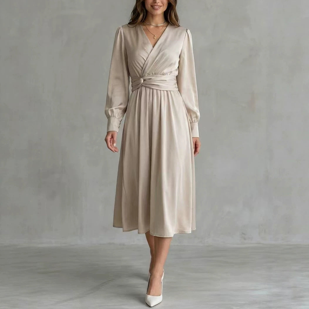 A woman models a light beige wrap midi dress with long sleeves, a matching fabric tie belt at the waist, and white pointed-toe heels against a textured gray studio backdrop.