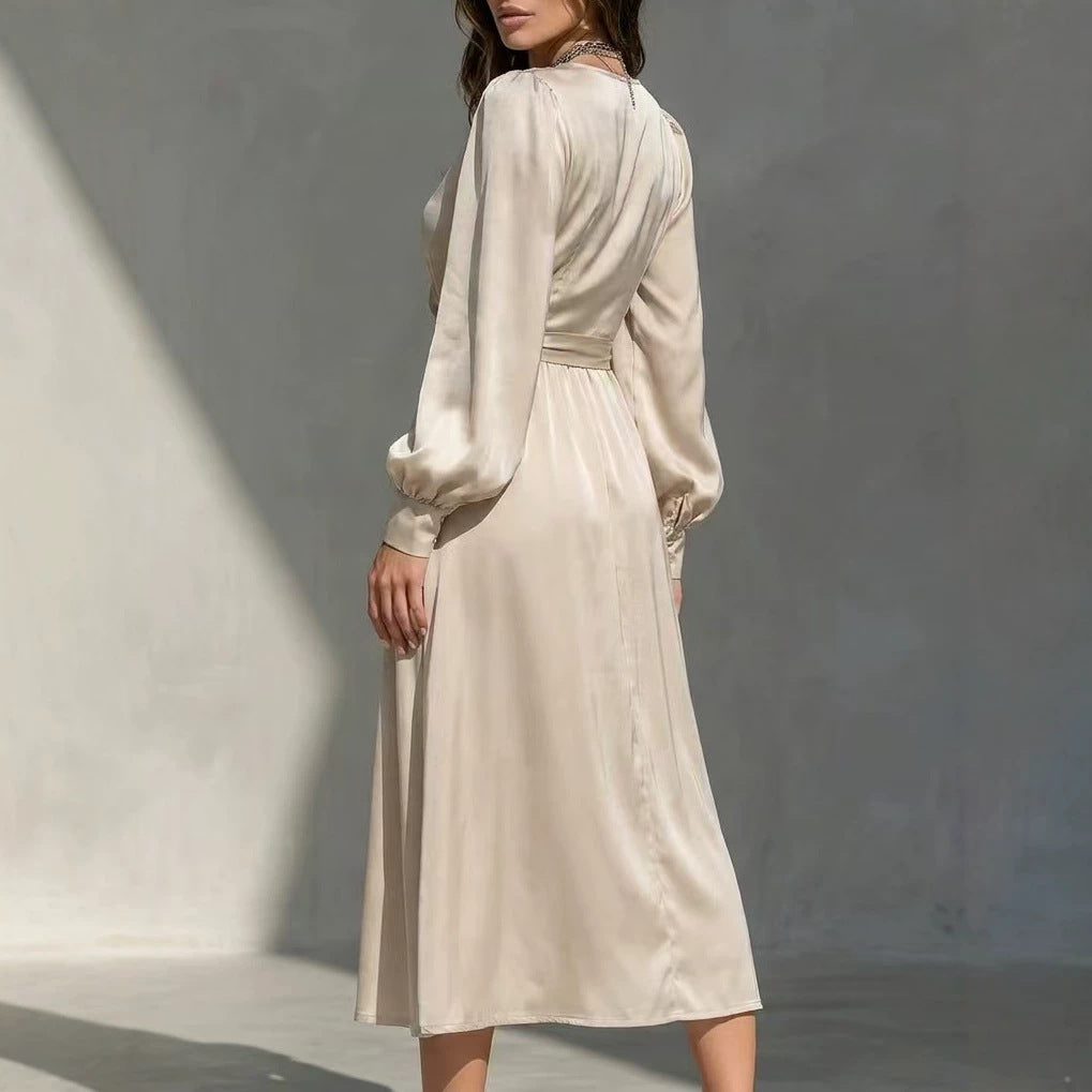 A woman wearing a long-sleeve midi dress in a light cream or beige satin-like fabric, photographed from the back against a textured gray wall with strong sunlight and shadows.