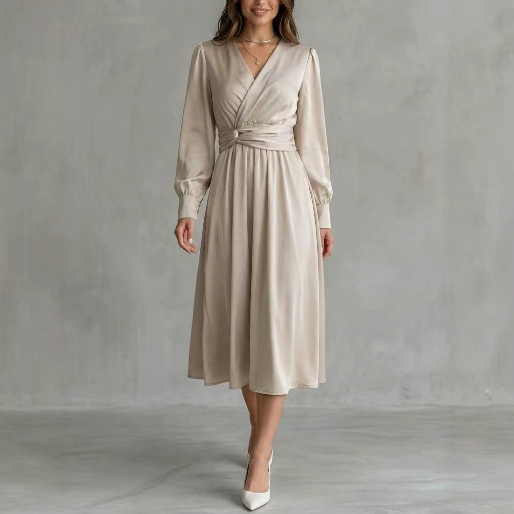 A woman models a light beige satin midi dress with a wrap-style V-neck, long sleeves, and a matching fabric tie belt at the waist, paired with white pointed-toe heels against a textured gray studio backdrop.