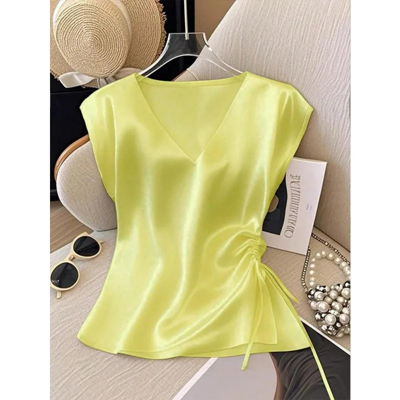 A light yellow-green satin top with a V-neckline, cap sleeves, and a side ruching detail with a tie, displayed on a hanger in a styled flat-lay setting.