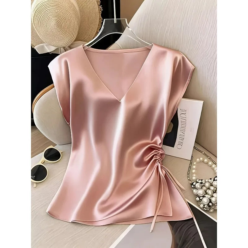 A satin-like blush pink blouse with a V-neckline, short sleeves, and a side drawstring detail, styled on a hanger in a tabletop flat-lay setting with a hat, sunglasses, card, and pearl accessories nearby.