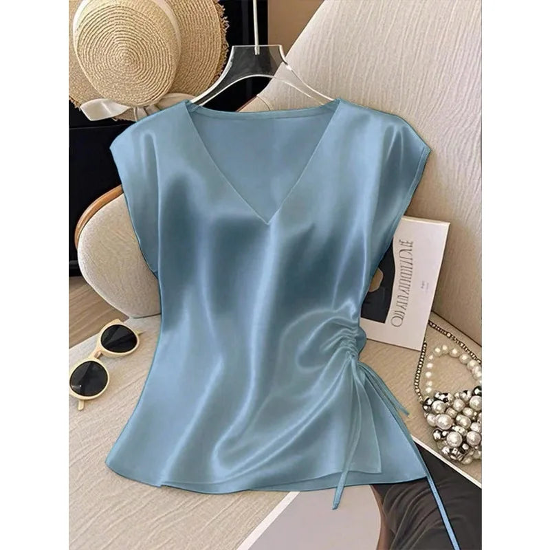 A light blue satin V-neck top with cap sleeves and a side drawstring, displayed on a hanger in a styled flat-lay setting with a straw hat, sunglasses, pearl necklace, and book.