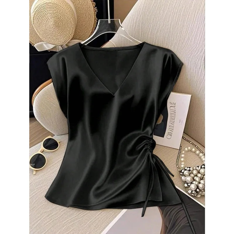 A black satin short-sleeve top with a V-neckline and side twist detail, displayed on a hanger in a styled flat-lay setting with a straw hat, sunglasses, pearls, and a book.