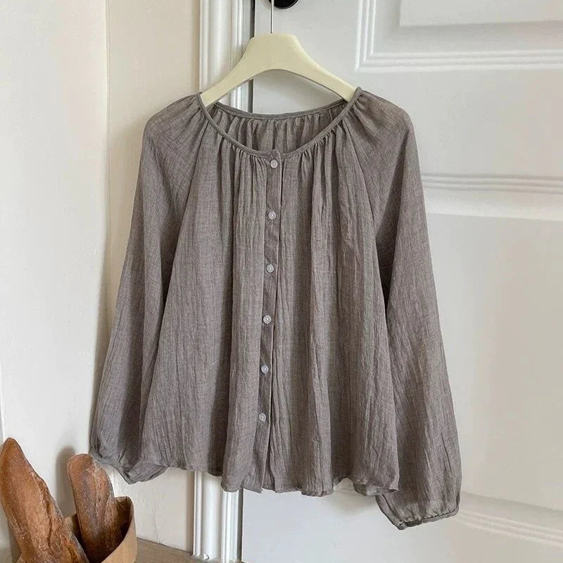 A taupe gray crinkle gauze blouse with a button-down front and gathered neckline, hanging on a cream hanger against a white paneled door.