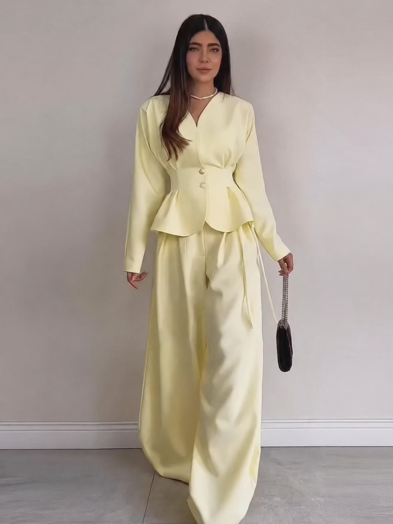 A woman modeling a pale yellow two-piece outfit consisting of a long-sleeve belted jacket and matching wide-leg trousers, standing against a plain light gray wall.