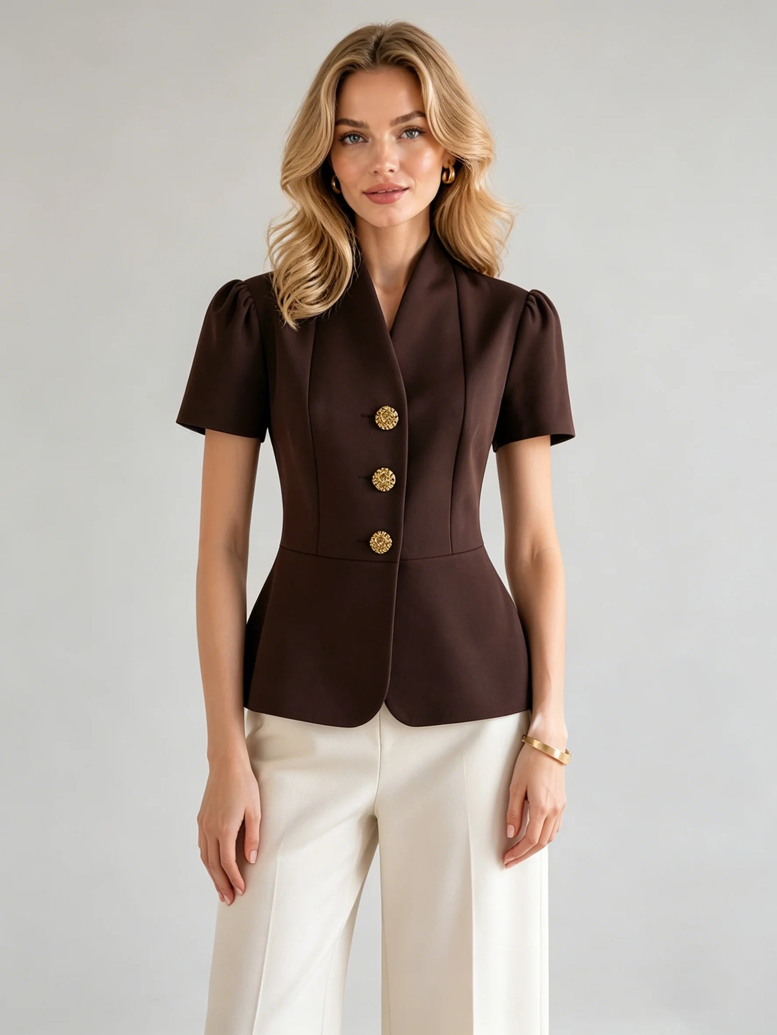 A woman modeling a dark brown short-sleeve blazer with gold buttons, paired with off-white trousers, against a light gray studio background.