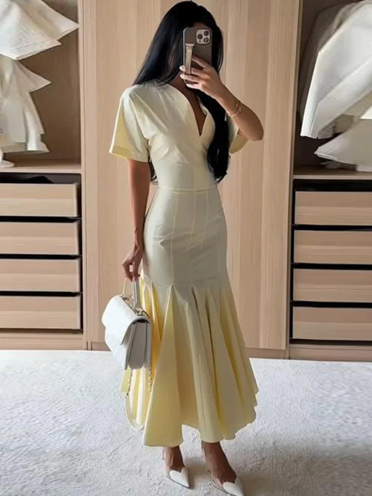 A woman takes a mirror selfie in a fitting room wearing a pale yellow dress with short sleeves, a deep V-neckline, and a flared mermaid-style hem. She is holding a white handbag and wearing white pointed shoes.