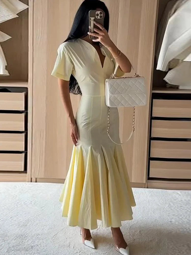 A woman takes a mirror selfie in a fitting room wearing a pale yellow short-sleeve mermaid-style dress with a deep V-neckline. She is holding a white quilted handbag with a gold chain strap and wearing white pointed-toe heels.
