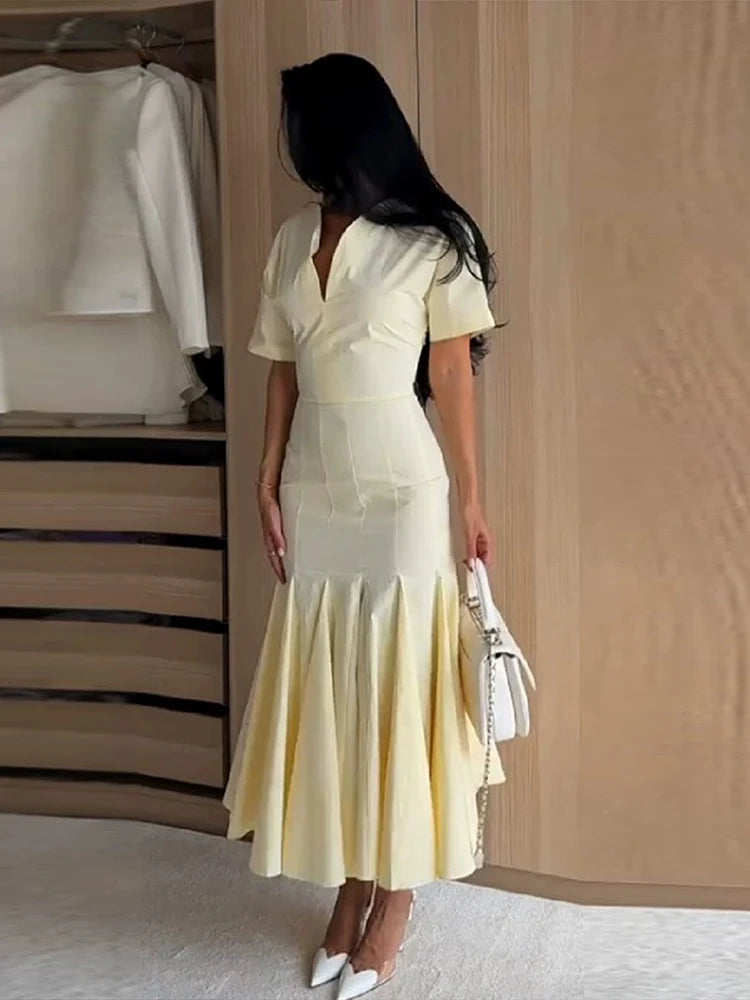 A woman stands in a room wearing a pale yellow short-sleeve midi dress with a V-neckline and a flared mermaid-style skirt. She is holding a white handbag and wearing white pointed shoes.