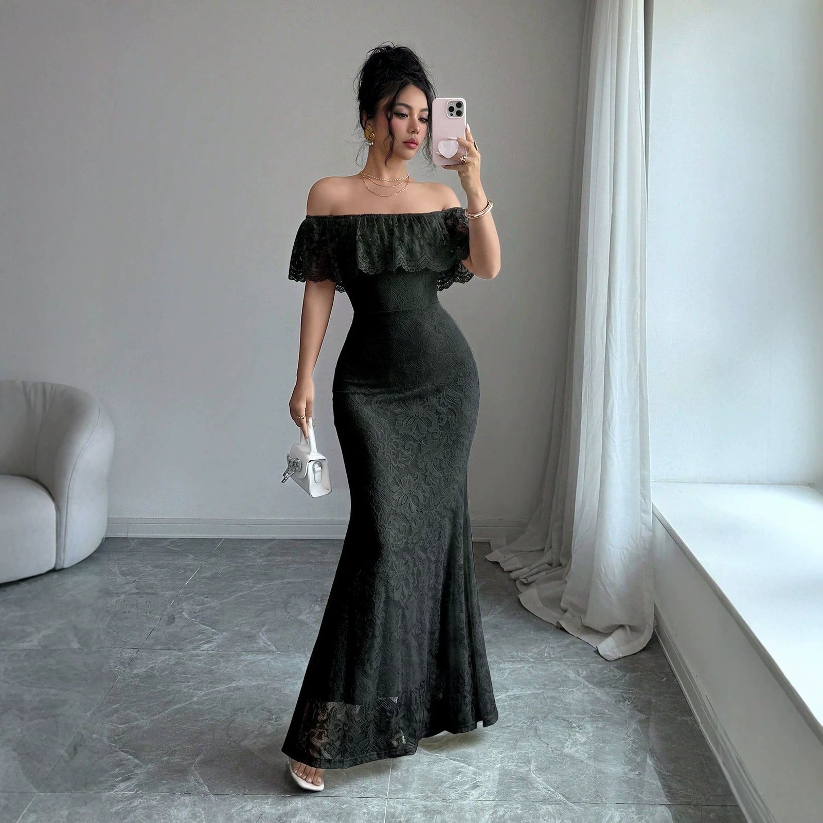 A woman takes a mirror selfie wearing a black off-the-shoulder lace maxi dress with a fitted silhouette and flared hem, holding a small white handbag in a bright room with gray marble flooring and white curtains.