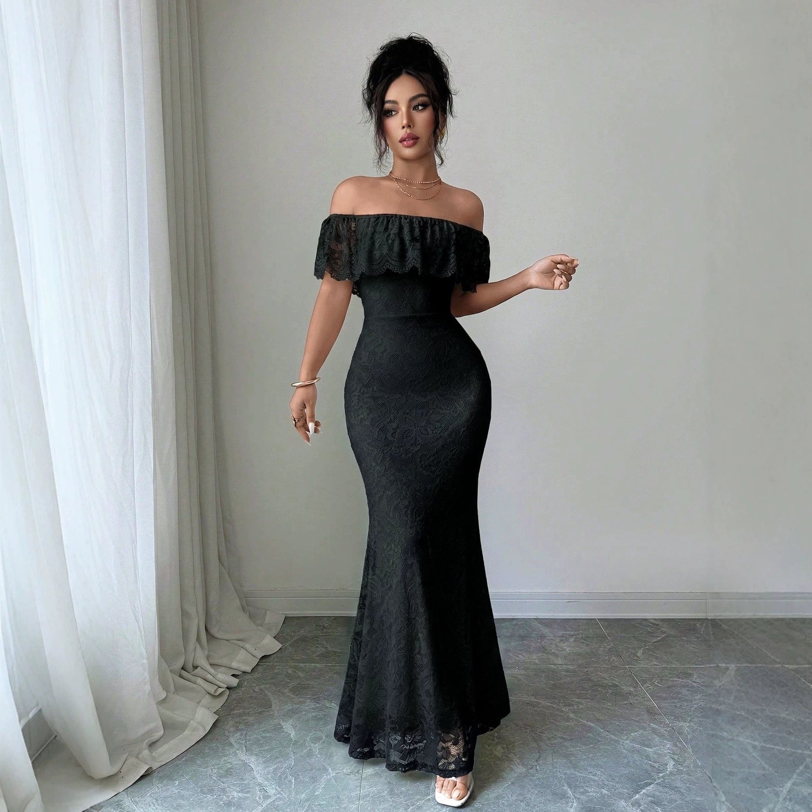 A woman models a black off-the-shoulder mermaid maxi dress with lace detailing, standing in a room with a gray marble floor and white curtains.