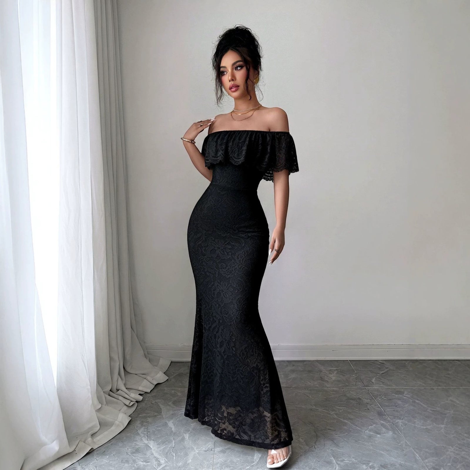 A woman models a black off-the-shoulder lace maxi dress in a minimalist room with white curtains and a gray floor.