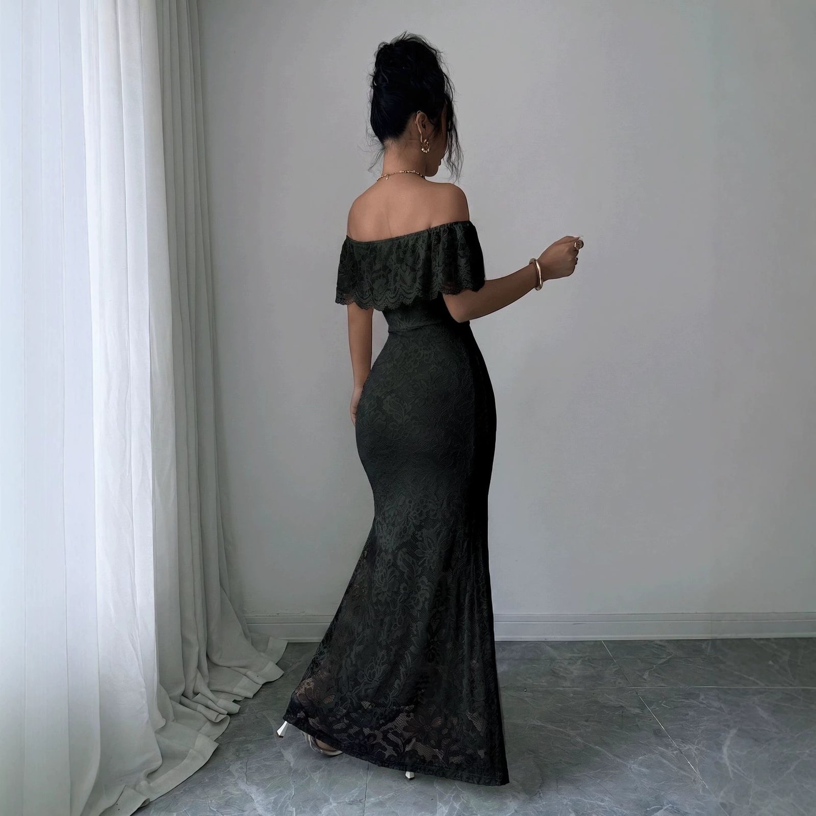 A woman wearing a black off-the-shoulder lace maxi dress, shown from the back in an indoor setting with a white curtain and gray tiled floor.