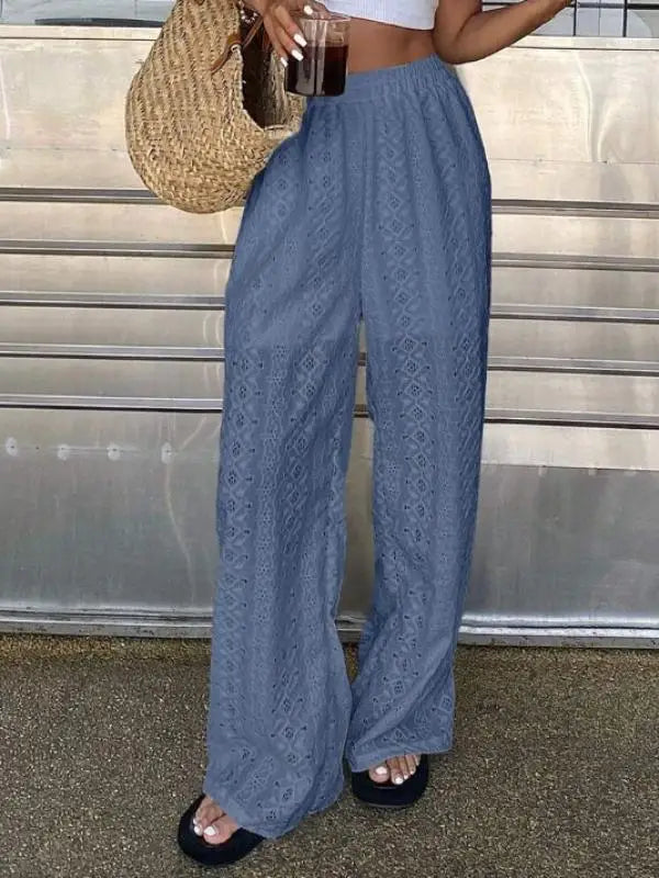 A person wearing loose, wide-leg blue pants with an allover textured eyelet or lace-like pattern, standing outdoors against a gray metal wall. The person is also holding a woven straw tote bag and a drink.
