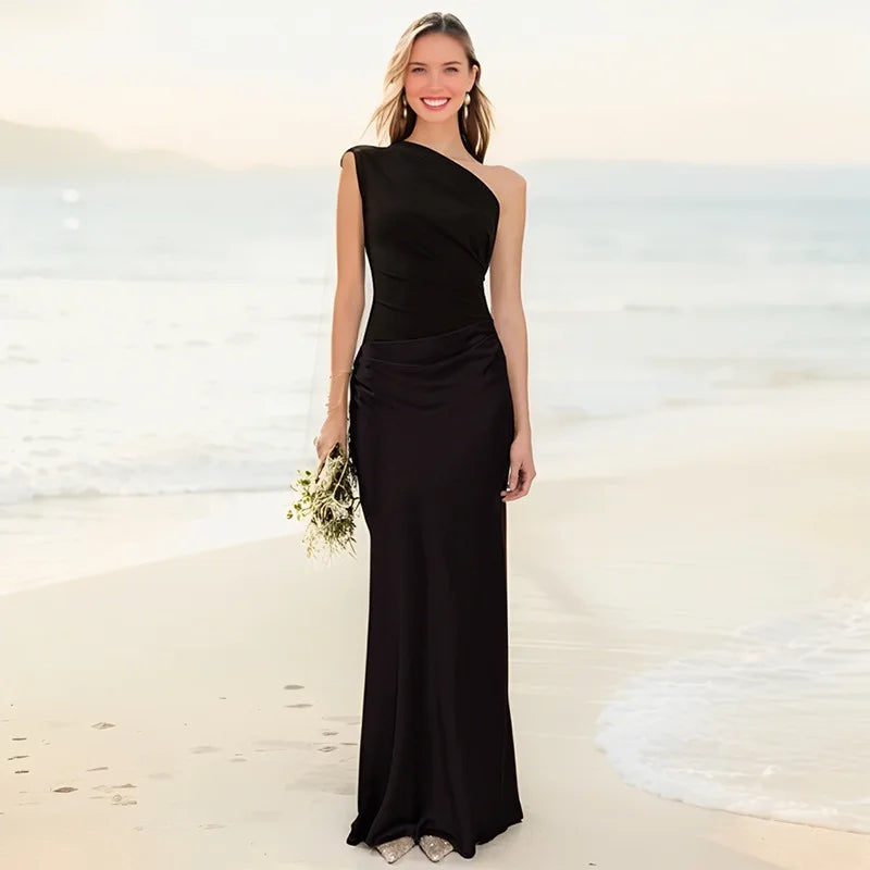 Woman in a black one-shoulder dress standing on a beach.