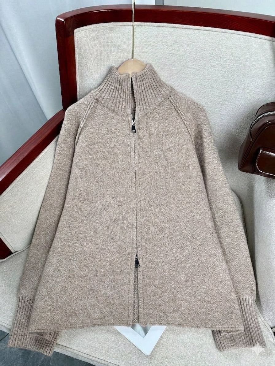 A beige knit zip-up sweater with a mock neck collar, displayed on a wooden hanger resting on a light beige upholstered chair.